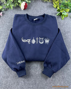Flat lay of a navy blue crewneck sweatshirt embroidered with gothic bookish symbols and Lady Death sleeve embroidery