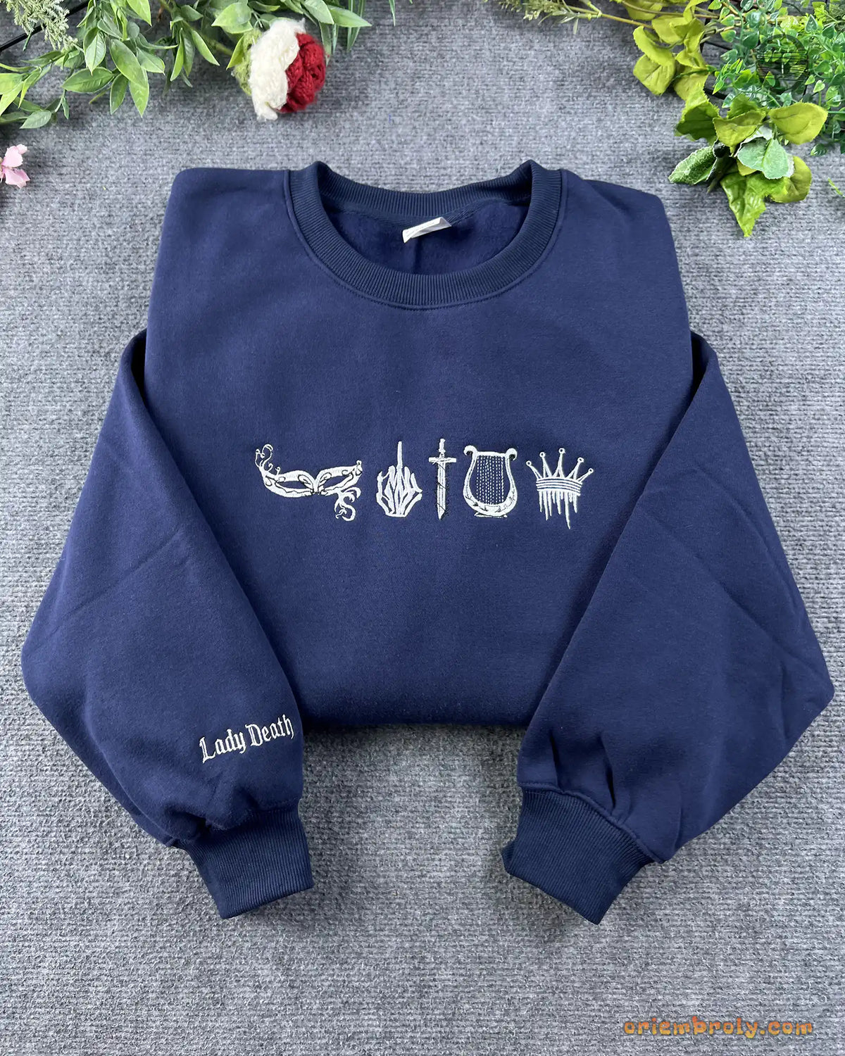 Flat lay of a navy blue crewneck sweatshirt embroidered with gothic bookish symbols and Lady Death sleeve embroidery