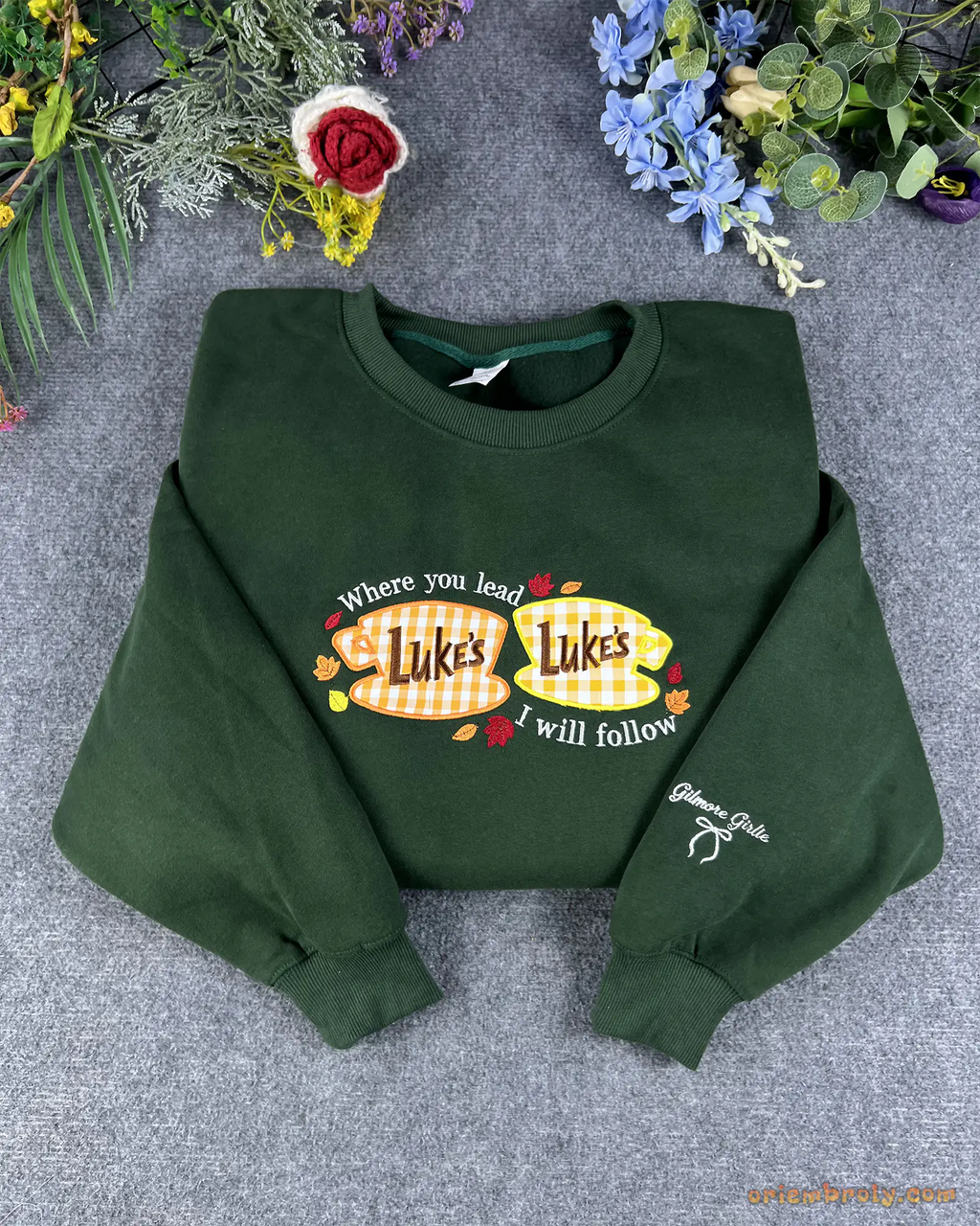 Flat lay of a forest green Gilmore Girls embroidered sweatshirt featuring Lukes Diner coffee cups and fall leaves