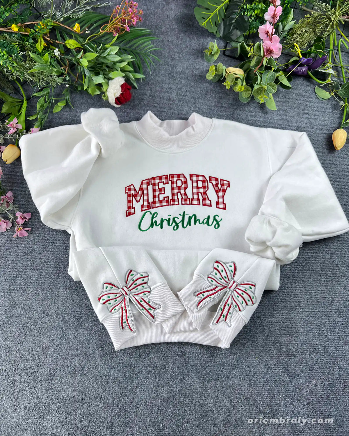 Flat lay of a white crewneck sweatshirt embroidered with red plaid Merry Christmas chest text and festive bow sleeve detail