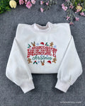 Flat lay of a white crewneck sweatshirt embroidered with red plaid Merry Christmas text and festive icons like reindeer and gingerbread