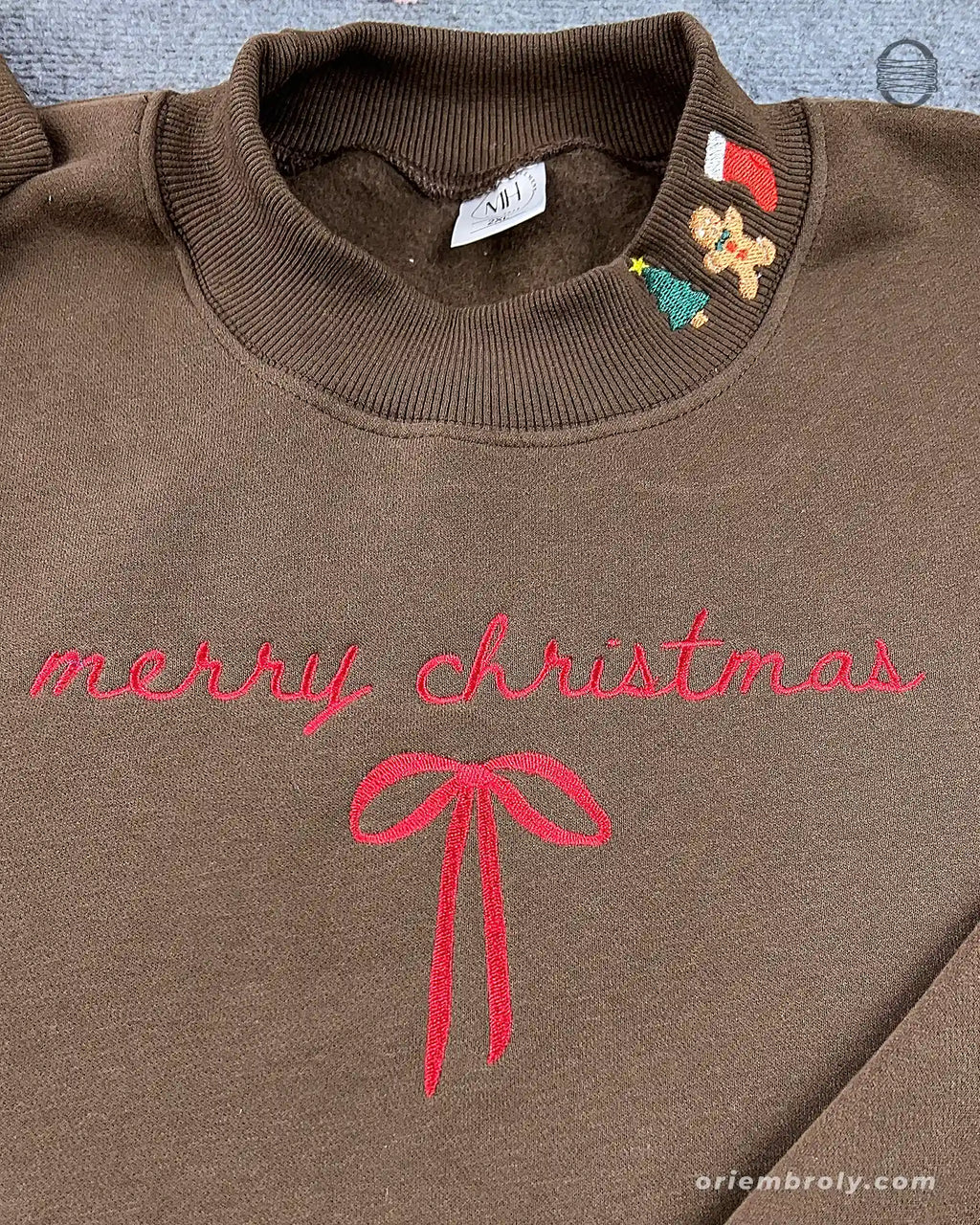 Close-up of red cursive Merry Christmas embroidery with ribbon bow and festive stocking gingerbread collar detail on a brown sweatshirt