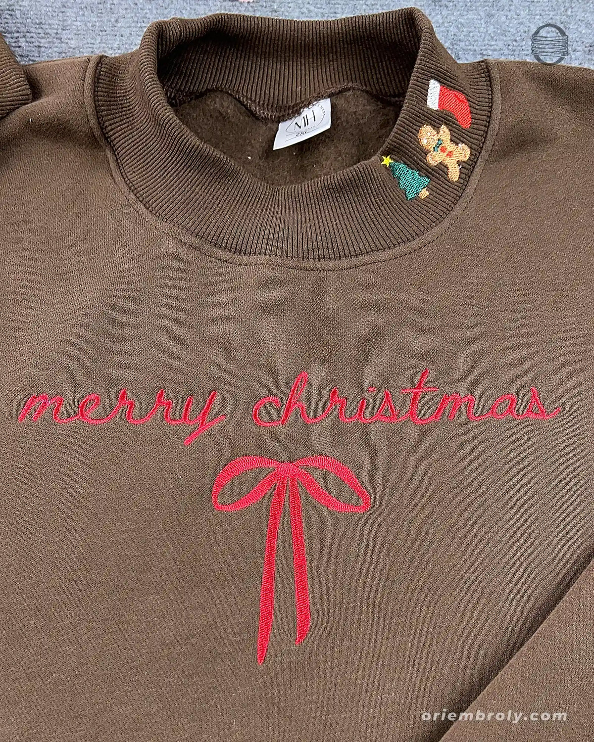 Close-up of red cursive Merry Christmas embroidery with ribbon bow and festive stocking gingerbread collar detail on a brown sweatshirt