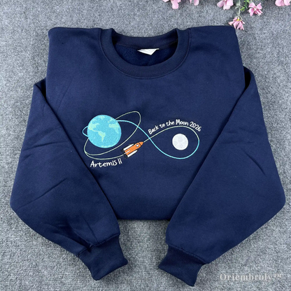 Premium navy blue crewneck sweatshirt featuring a detailed embroidered NASA Artemis II mission flight path, Earth, and Moon. High-quality space exploration apparel for fans
