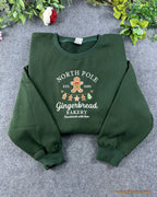 Flat lay of a forest green Christmas crewneck sweatshirt embroidered with North Pole Gingerbread Bakery and gingerbread man