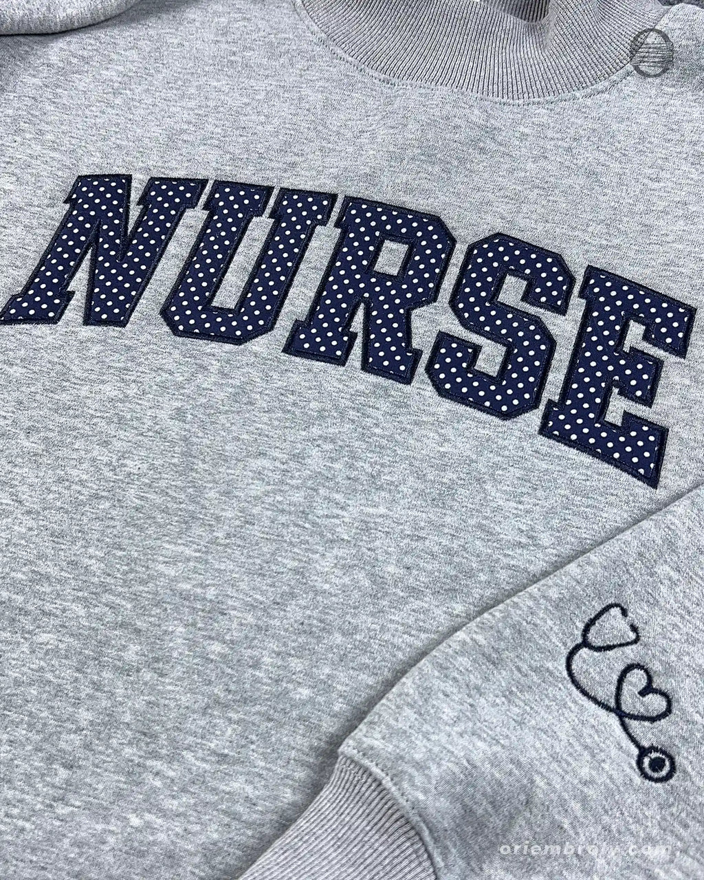 Close-up of textured dark varsity nurse chest embroidery and cute stethoscope with heart line embroidered on the sleeve of a grey sweater