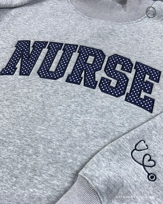 Close-up of textured dark varsity nurse chest embroidery and cute stethoscope with heart line embroidered on the sleeve of a grey sweater