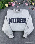 Flat lay of a sport grey crewneck sweatshirt with dark varsity nurse chest embroidery and stethoscope heart sleeve detail