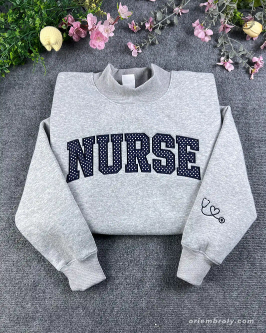Flat lay of a sport grey crewneck sweatshirt with dark varsity nurse chest embroidery and stethoscope heart sleeve detail