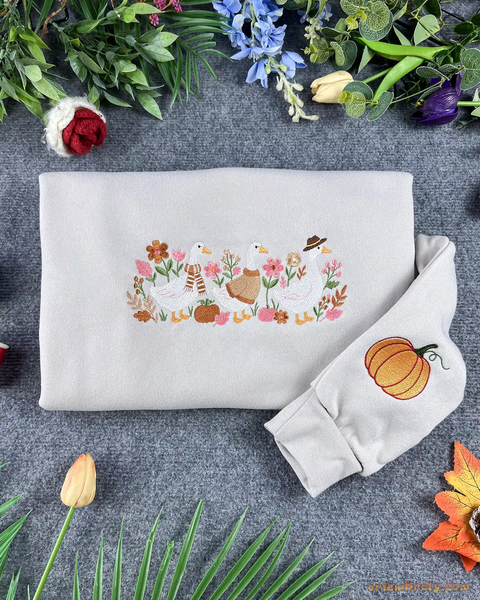 Flatlay view of an oatmeal beige crewneck sweatshirt embroidered with mama duck and ducklings surrounded by autumn flowers, with a pumpkin detail on the sleeve.