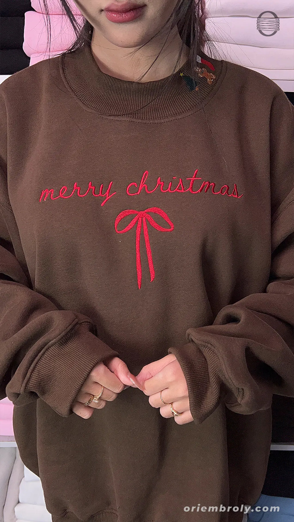 Model wearing an oversized chocolate brown Merry Christmas embroidered crewneck sweater with red ribbon bow indoors