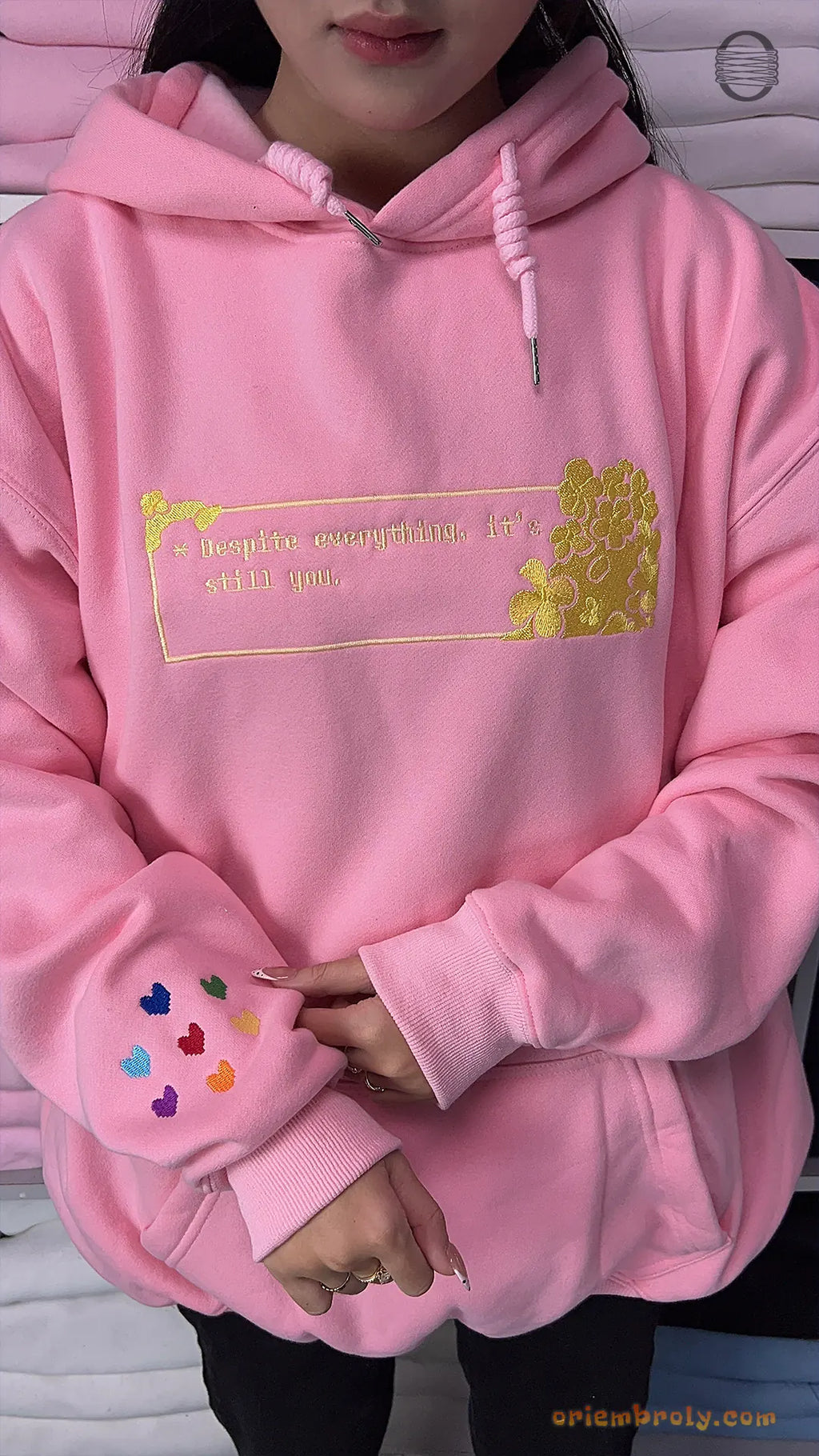 Model wearing an oversized light pink embroidered hoodie with gold romantic chest design and rainbow hearts sleeve detail