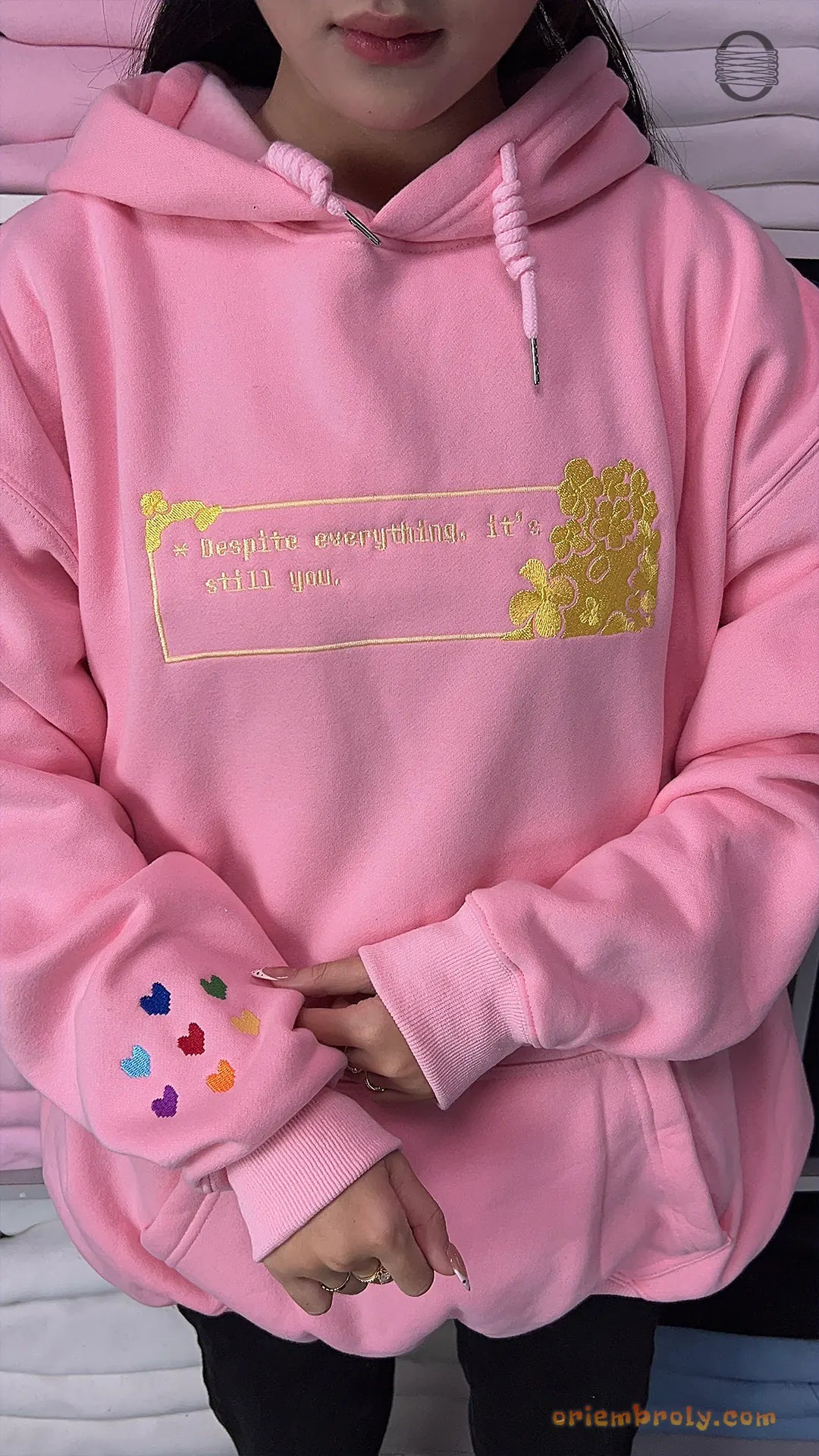 Model wearing an oversized light pink embroidered hoodie with gold romantic chest design and rainbow hearts sleeve detail
