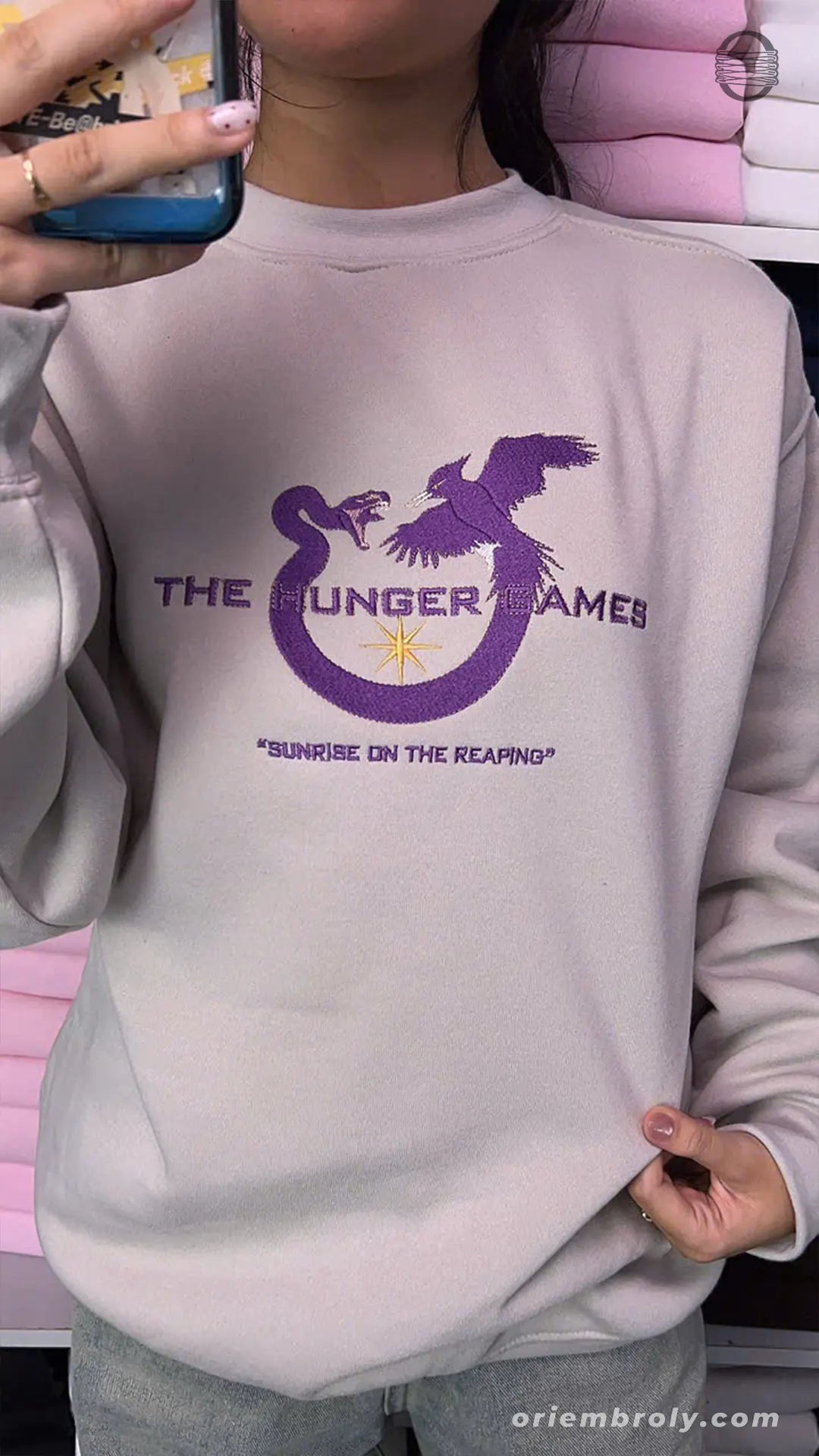 Model wearing an oversized sand crewneck sweater with purple Hunger Games Sunrise on the Reaping embroidery
