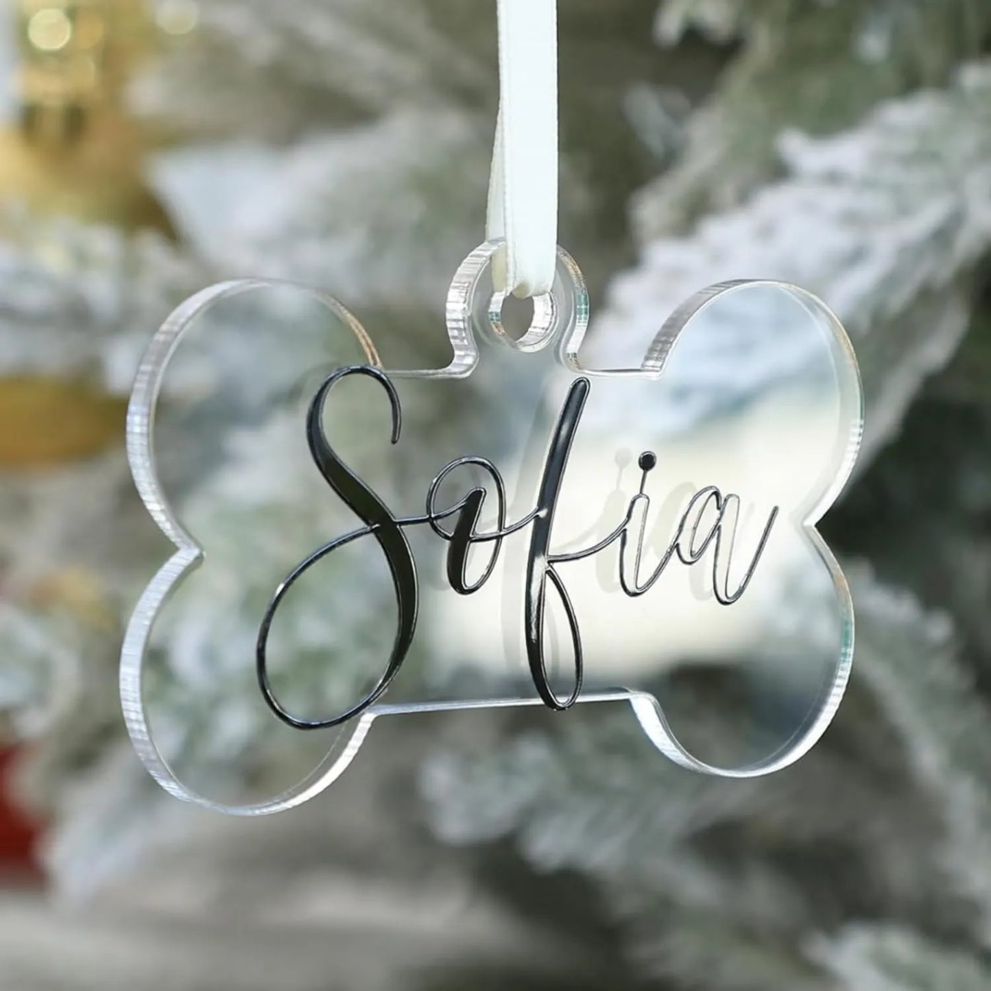 Personalized clear acrylic dog bone Christmas ornament with the name "Sofia" written in black script font, hanging from a white ribbon on a snowy Christmas tree.
