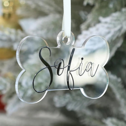 Personalized clear acrylic dog bone Christmas ornament with the name "Sofia" written in black script font, hanging from a white ribbon on a snowy Christmas tree.