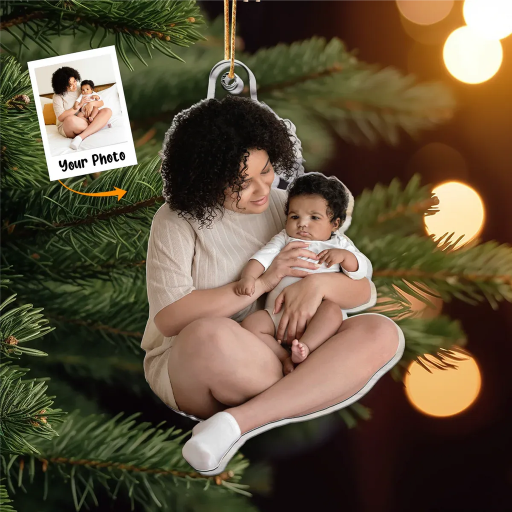 A personalized acrylic photo Christmas ornament featuring a mother holding her baby, designed as a keepsake for a new family's first Christmas. The custom ornament is hanging on a festive green Christmas tree with blurred warm lights in the background