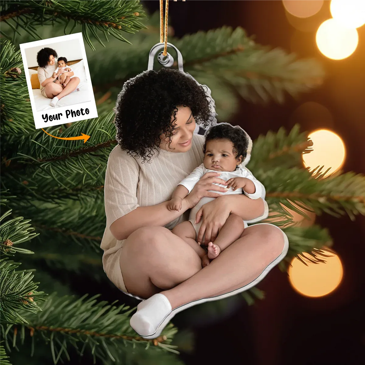 A personalized acrylic photo Christmas ornament featuring a mother holding her baby, designed as a keepsake for a new family's first Christmas. The custom ornament is hanging on a festive green Christmas tree with blurred warm lights in the background