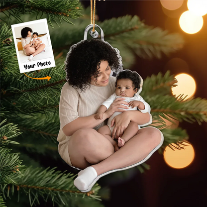 A personalized acrylic photo Christmas ornament featuring a mother holding her baby, designed as a keepsake for a new family's first Christmas. The custom ornament is hanging on a festive green Christmas tree with blurred warm lights in the background