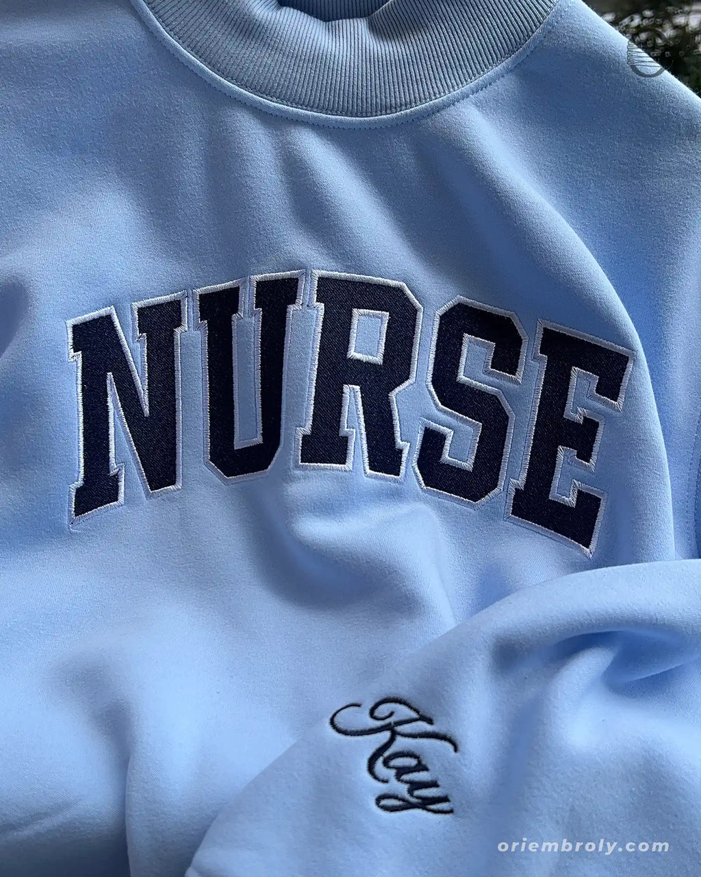 Close-up of black varsity nurse chest embroidery and custom handwritten name script on the sleeve of a light blue sweatshirt