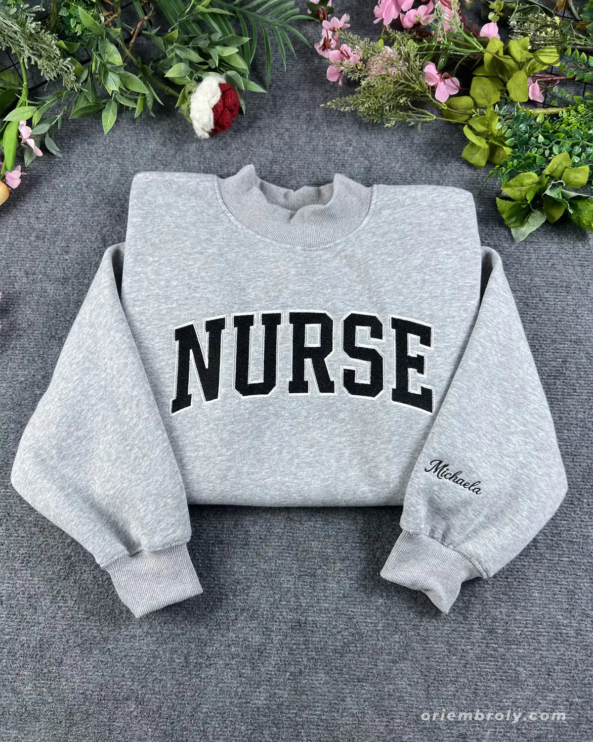 Flat lay of a sport grey sweatshirt embroidered with black varsity nurse block letters and custom name sleeve detail