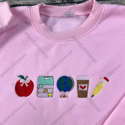 Pink sweatshirt with embroidered icons including an apple, book, world, coffee cup, and pencil.