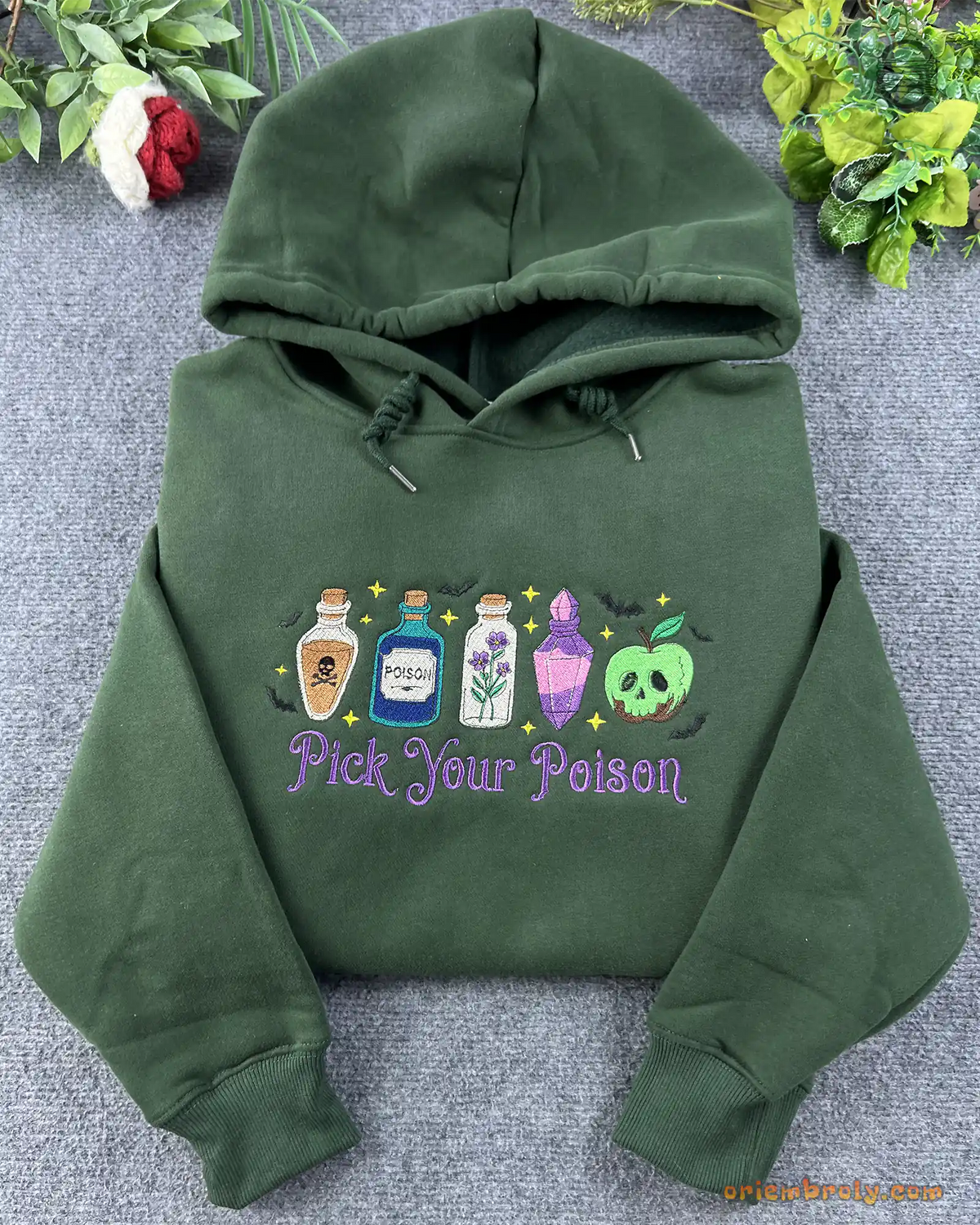 Flat lay of a forest green Halloween hoodie embroidered with Pick Your Poison and colorful witch potion bottles