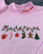 Close-up of red Merry Christmas collar embroidery and festive garland featuring gingerbread man, stocking, and Christmas tree on a pink sweater