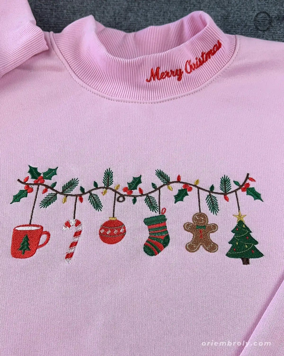 Close-up of red Merry Christmas collar embroidery and festive garland featuring gingerbread man, stocking, and Christmas tree on a pink sweater