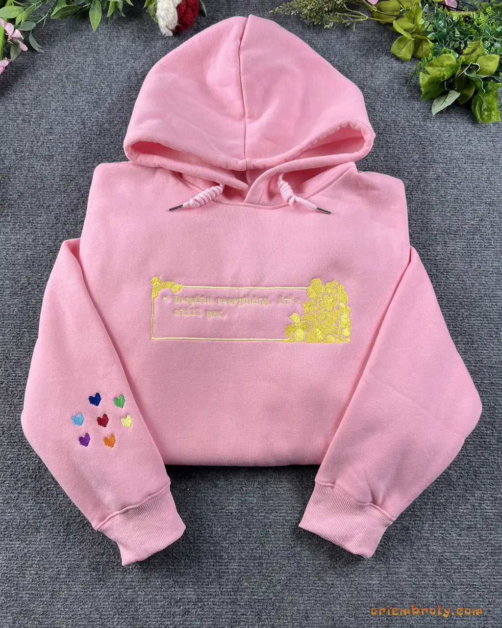Flat lay of a light pink hoodie embroidered with gold romantic hearts quote and colorful rainbow hearts on sleeve