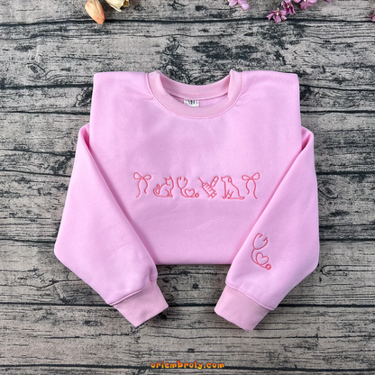 Pink embroidered sweatshirt featuring cat, dog, syringe, stethoscope, and heart designs — perfect for veterinarians or pet lovers.