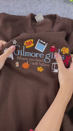 Video close-up showing Gilmore Girls embroidered text and coffee cup details on a brown crewneck sweatshirt