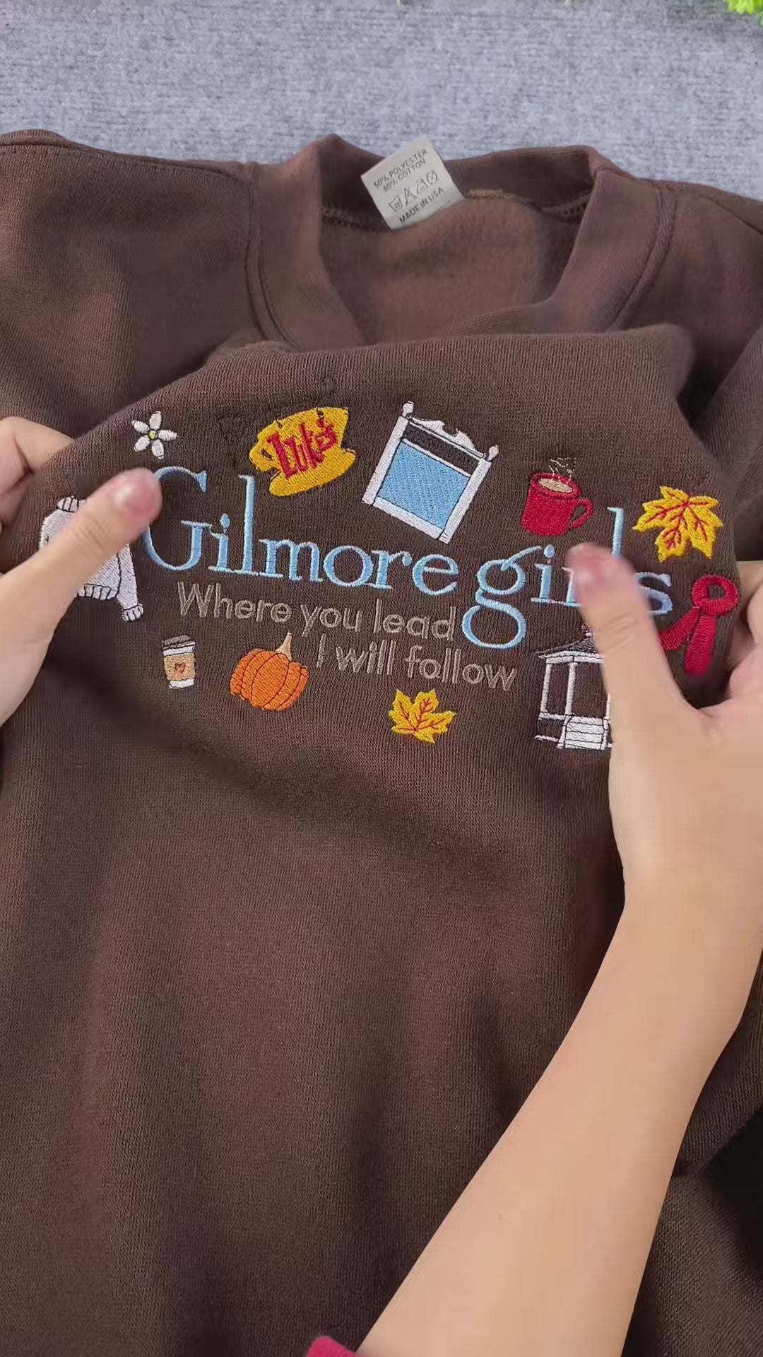 Video close-up showing Gilmore Girls embroidered text and coffee cup details on a brown crewneck sweatshirt