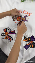 Close-up video screenshot of a person carefully cutting thread from a detailed Halloween Halloweentown embroidery design on a white crewneck sweatshirt, featuring pumpkin and witch hat motifs with large decorative bows below.