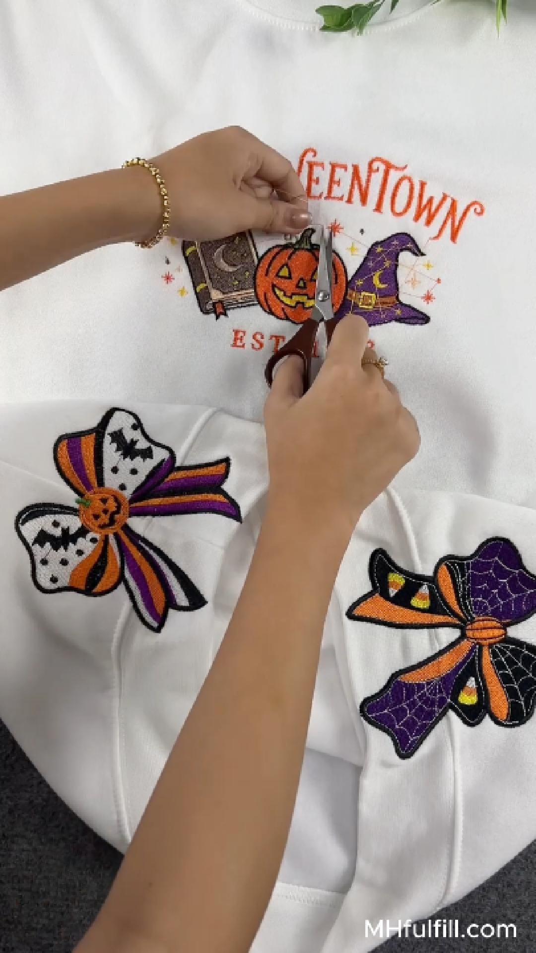 Close-up video screenshot of a person carefully cutting thread from a detailed Halloween Halloweentown embroidery design on a white crewneck sweatshirt, featuring pumpkin and witch hat motifs with large decorative bows below.