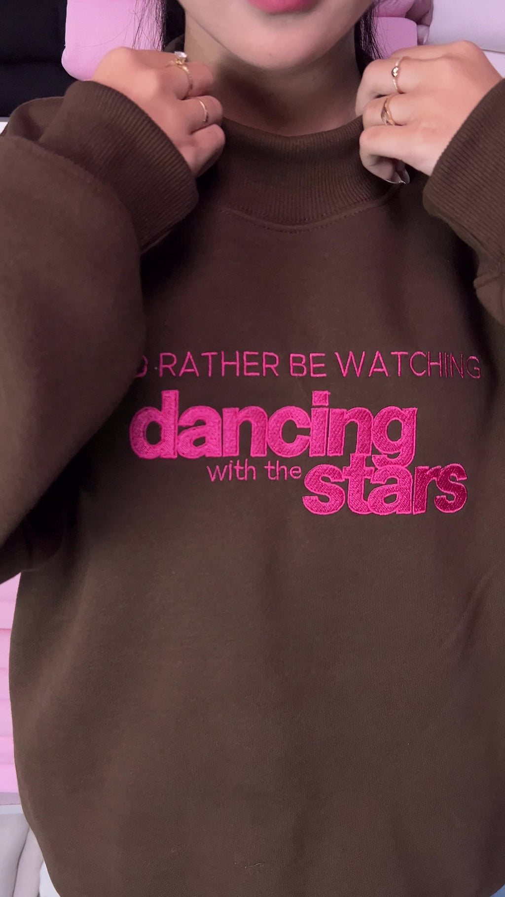 Video showing vibrant hot pink embroidery detail of I'd Rather Be Watching Dancing with the Stars on a brown sweatshirt
