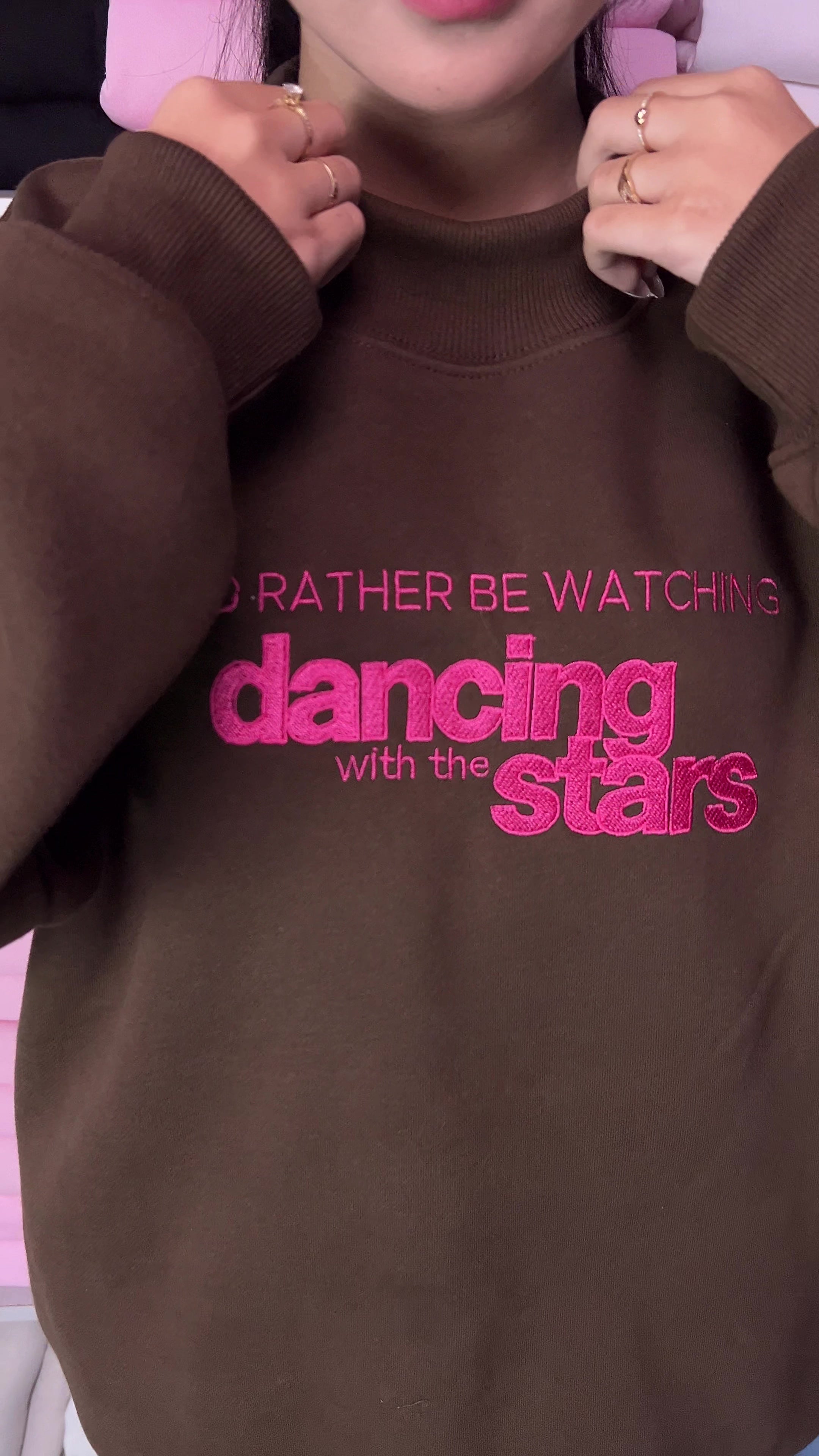 Video showing vibrant hot pink embroidery detail of I'd Rather Be Watching Dancing with the Stars on a brown sweatshirt