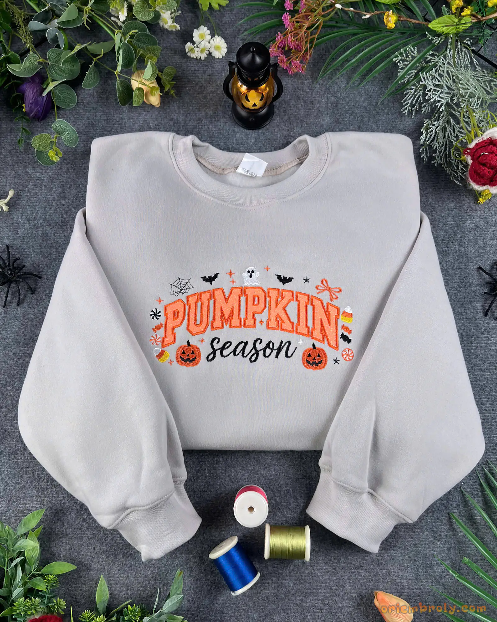 Flat lay of a light grey crewneck sweatshirt embroidered with orange Pumpkin Season text, pumpkins, bats, and ghost details