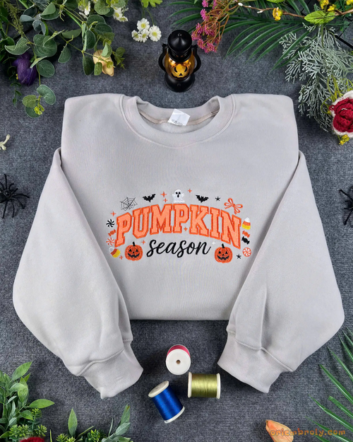Flat lay of a light grey crewneck sweatshirt embroidered with orange Pumpkin Season text, pumpkins, bats, and ghost details