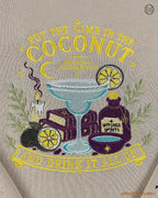 Close-up of yellow Put The Lime In The Coconut embroidered graphics on a beige sweatshirt