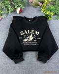 Flat lay of a black crewneck sweatshirt embroidered with Salem Massachusetts Est 1626 vintage flying witch design