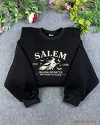 Flat lay of a black crewneck sweatshirt embroidered with Salem Massachusetts Est 1626 vintage flying witch design