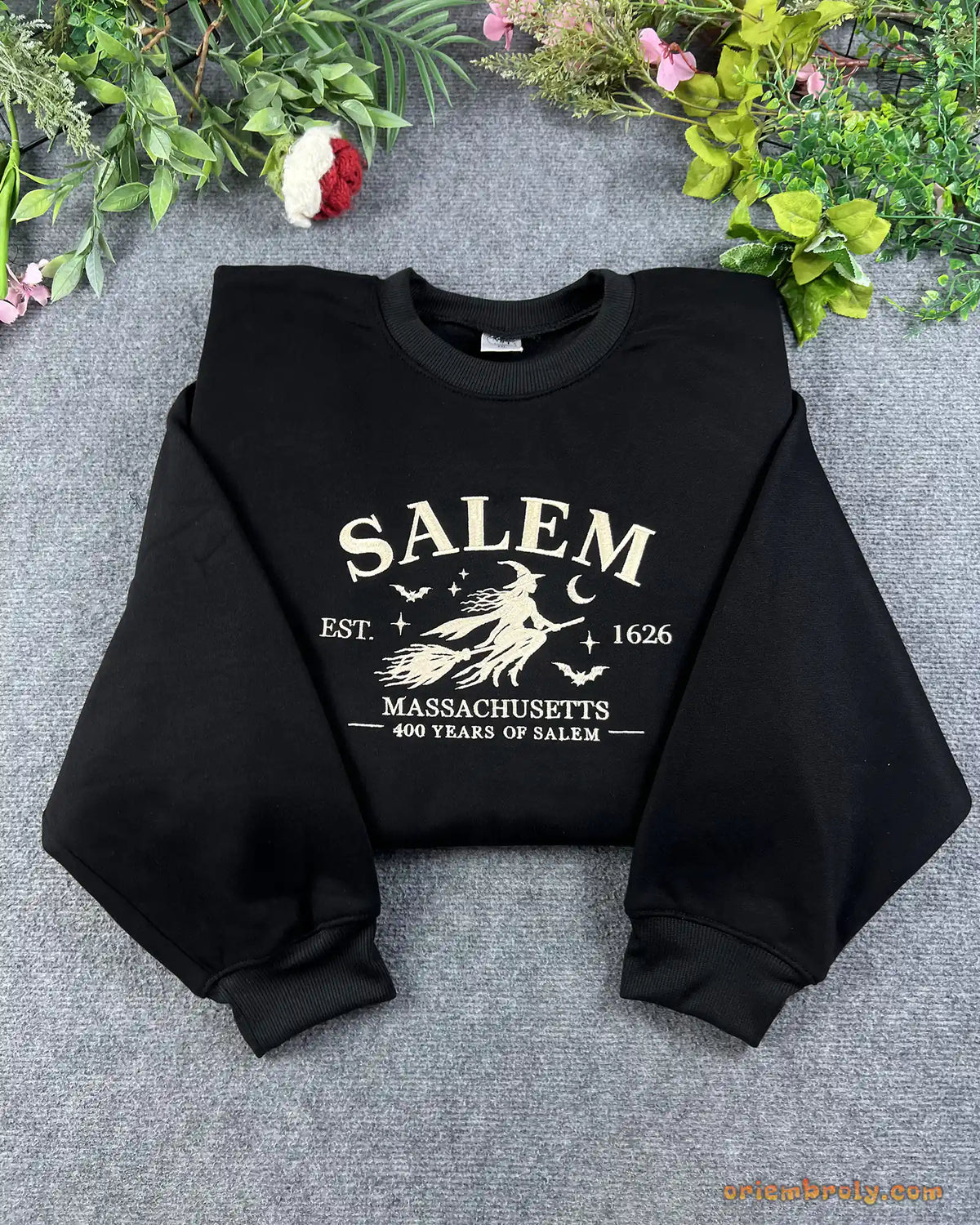 Flat lay of a black crewneck sweatshirt embroidered with Salem Massachusetts Est 1626 vintage flying witch design