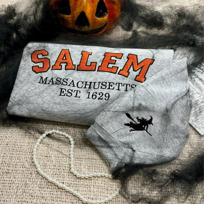 Gray face mask with 'Salem Massachusetts Est. 1629' text and witch design, Halloween pumpkin in background.