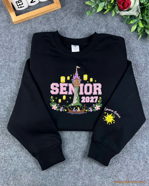 Flat lay of a black Senior 2027 sweatshirt featuring Tangled Rapunzel tower, floating lanterns, and sun emblem sleeve embroidery