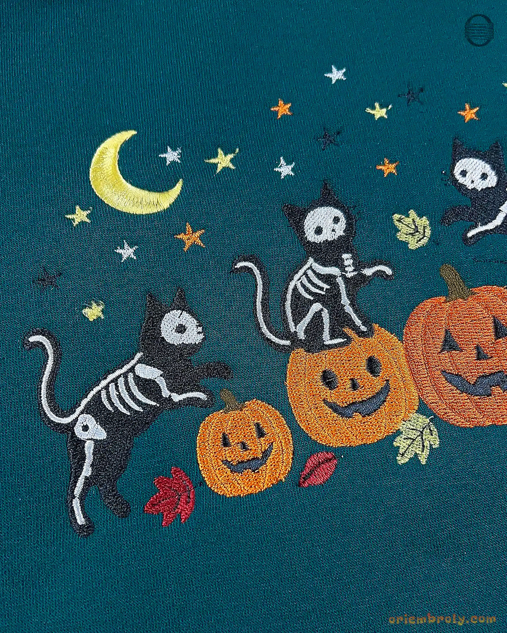 Close-up of embroidered skeleton cats sitting on jack-o-lantern pumpkins with yellow stars and autumn leaves.