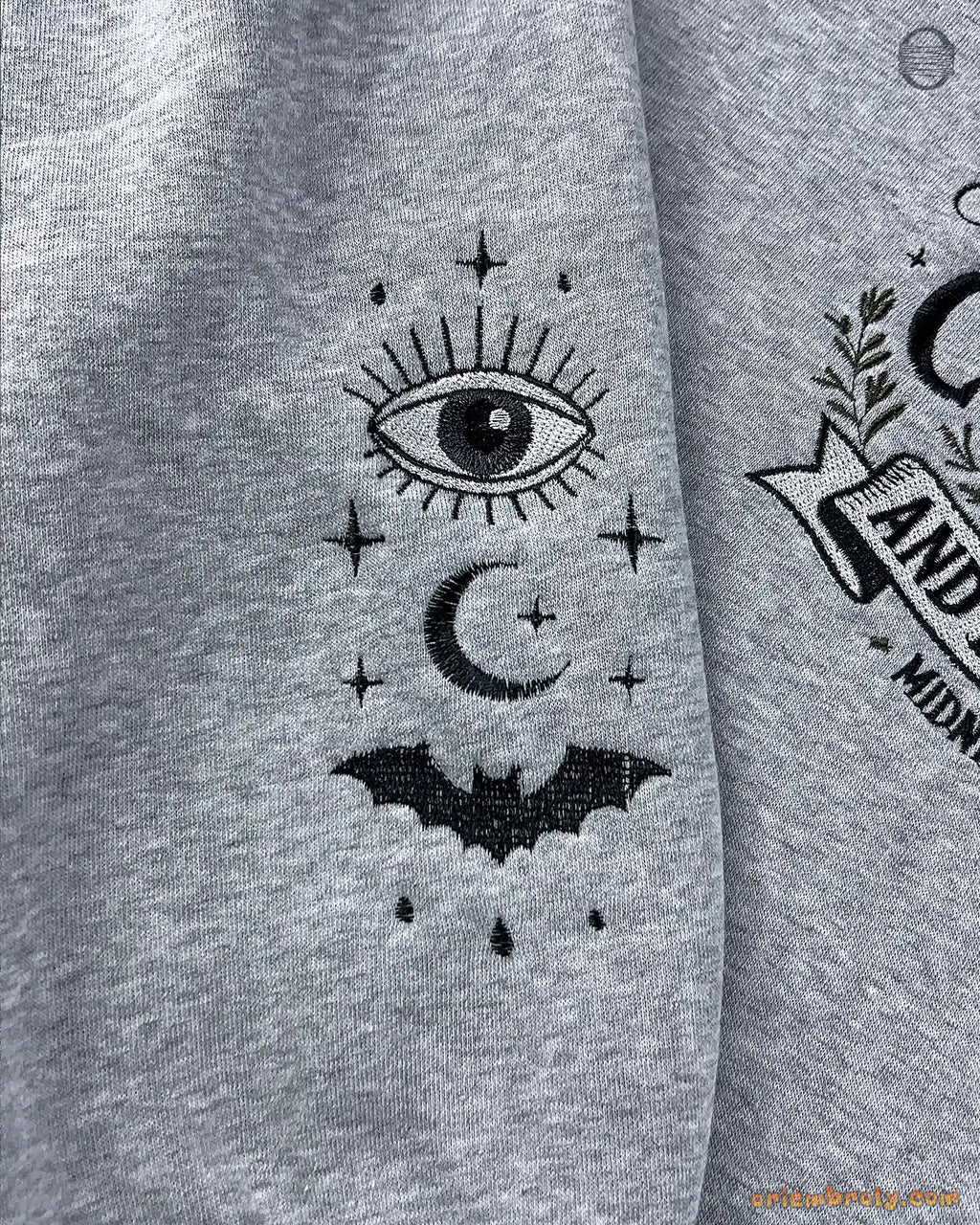Close-up of celestial sleeve embroidery featuring mystical eye, crescent moon, and bat on a grey sweatshirt