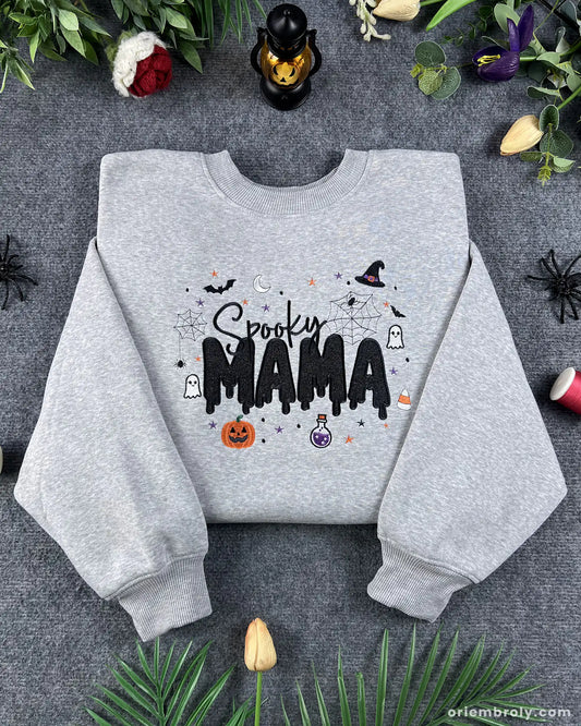Flatlay view of an ash gray crewneck sweatshirt embroidered with Spooky MAMA text, pumpkin, witch hat, potion bottle, and bats.