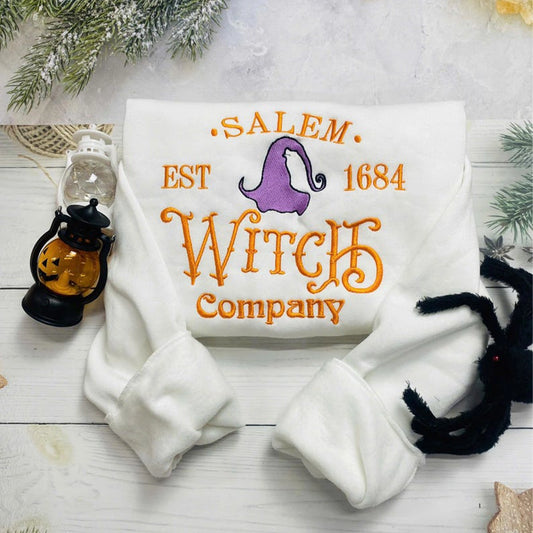 White pillow with 'Salem Est 1684 Witch Company' text and purple witch hat design on a wooden surface with Halloween decorations.