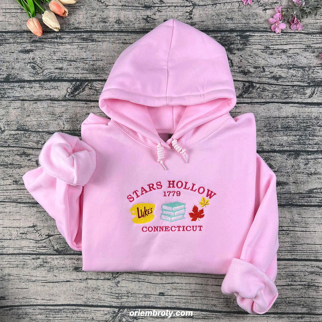 Pink hoodie with 'Stars Hollow 1779 Loves Connecticut' text and graphics on a wooden surface.